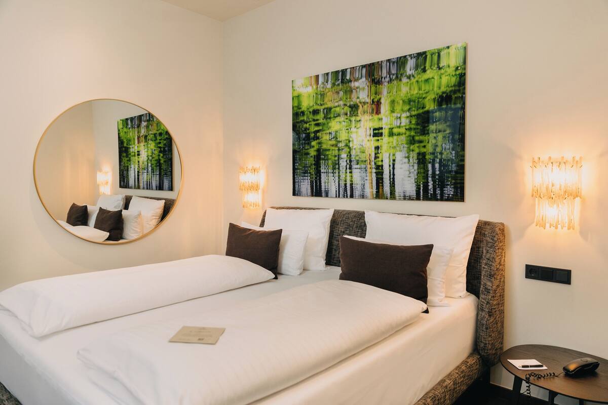Un letto matrimoniale con biancheria da letto bianco e marrone, uno specchio rotondo, un quadro raffigurante alberi e due applique.