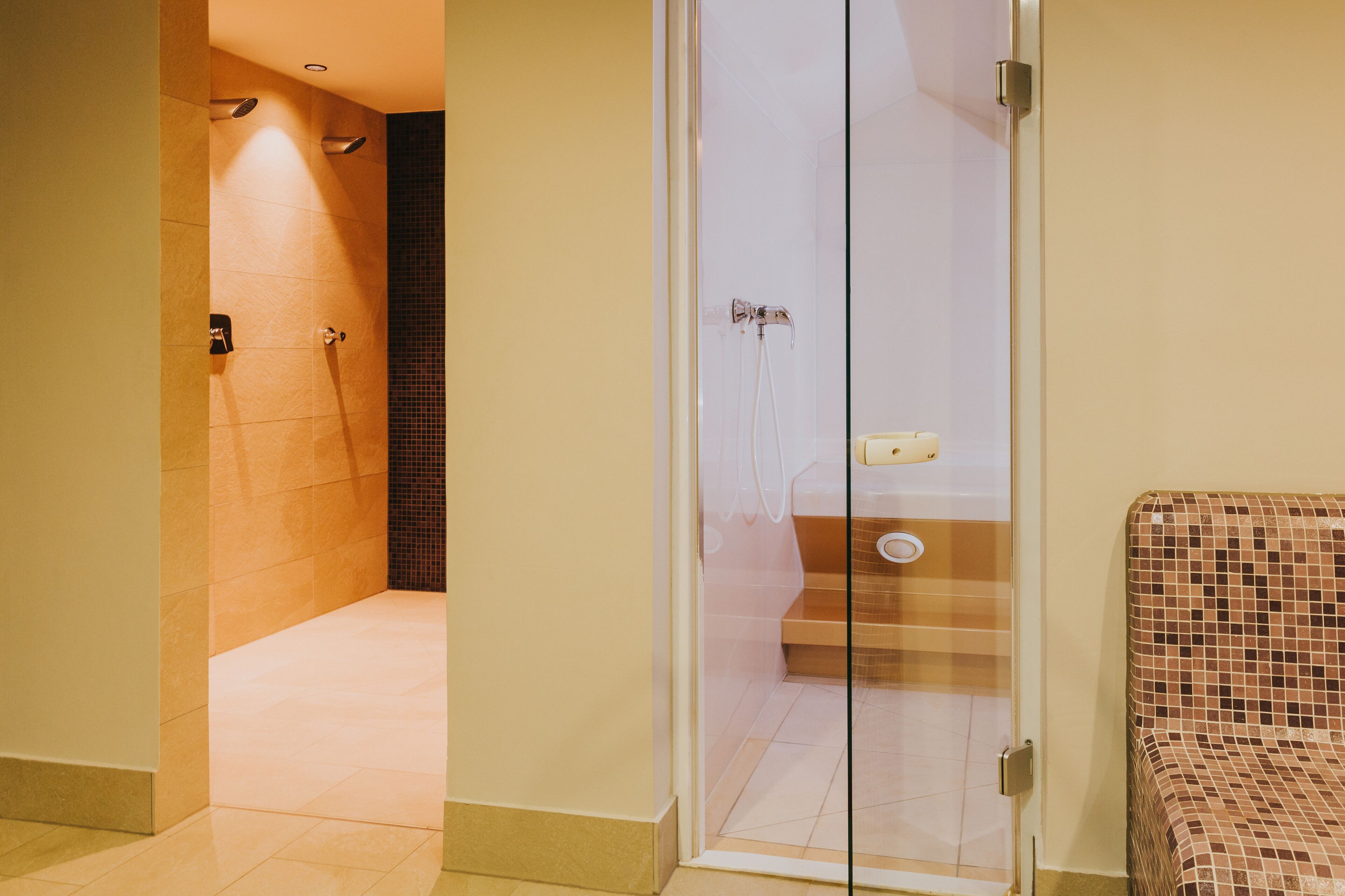 A hotel bathroom with a glass shower enclosure, a tiled floor, and a small seating area with a patterned seat.