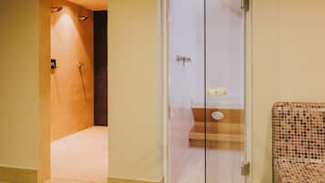 A hotel bathroom with a glass shower enclosure, a tiled floor, and a small seating area with a patterned seat.