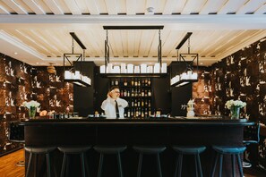 Uma área de bar com um bartender, um balcão, banquetas e uma parede com garrafas de bebidas.