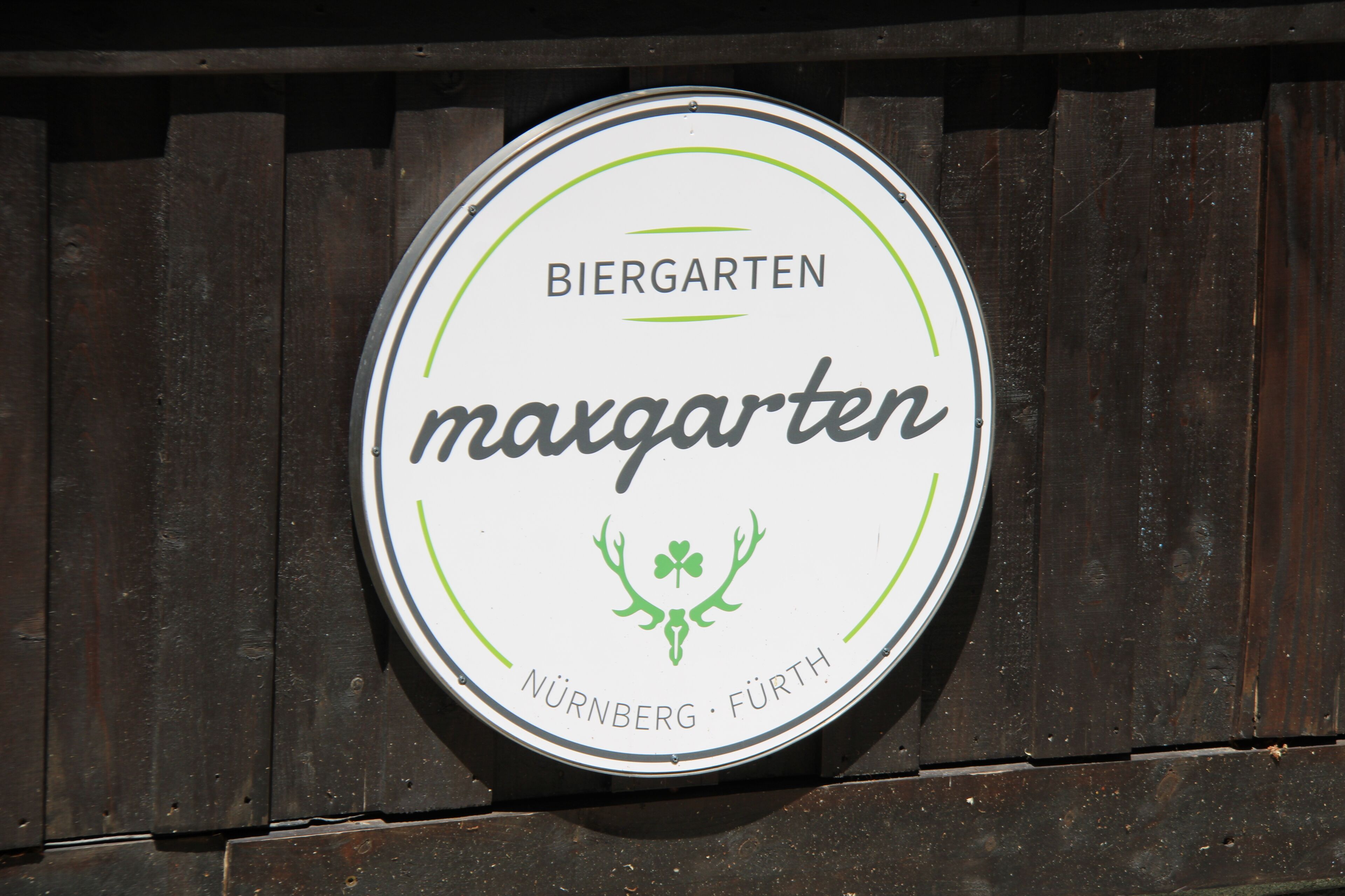 Ein rundes Schild mit dem Text 'BIERGARTEN maxgarten' und einer kleinen Darstellung eines Kleeblatts.