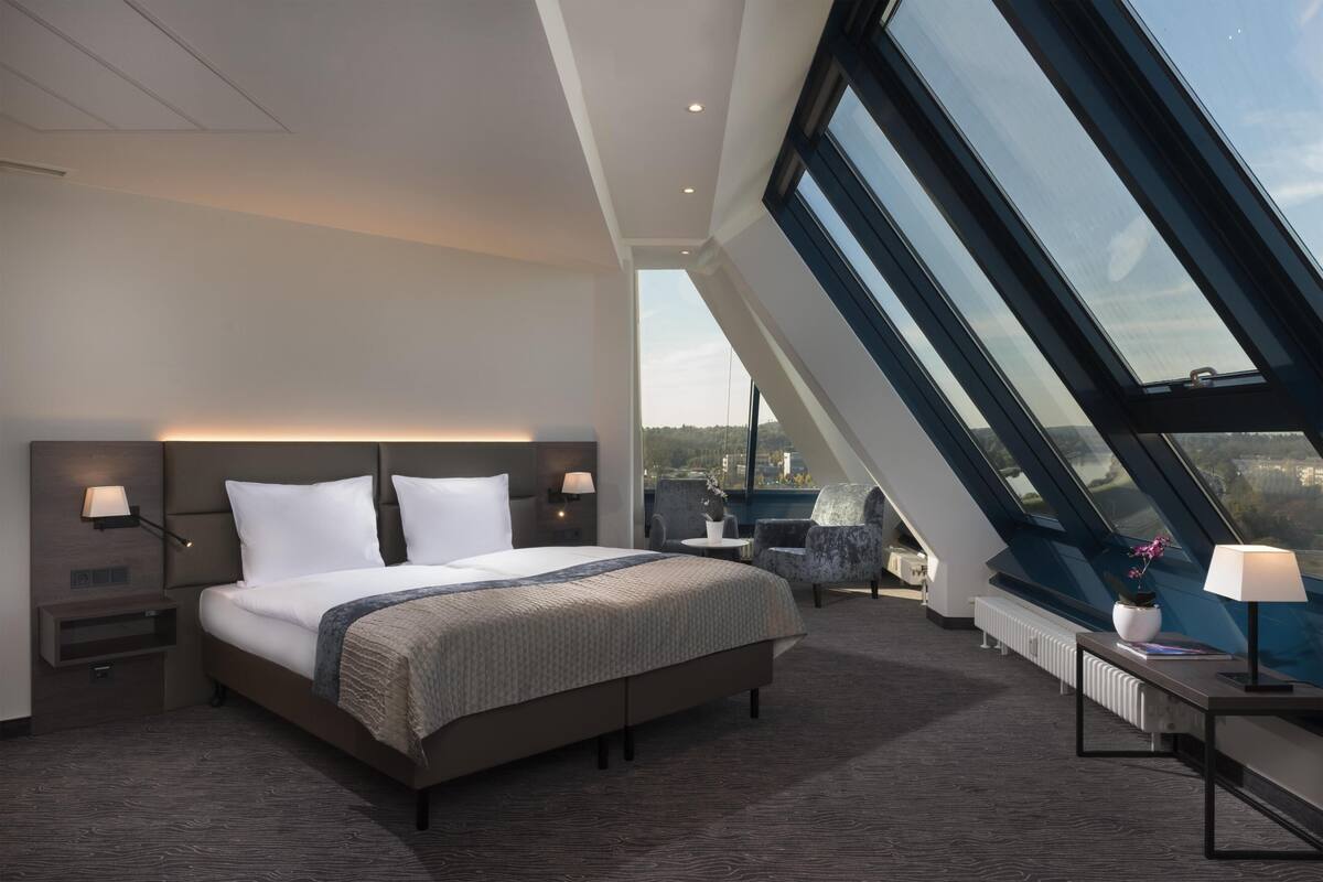 Ein modernes Hotelzimmer mit einem großen Bett, einem Schreibtisch, einem Sessel und Blick auf die Stadt, inklusive Dachfenster.