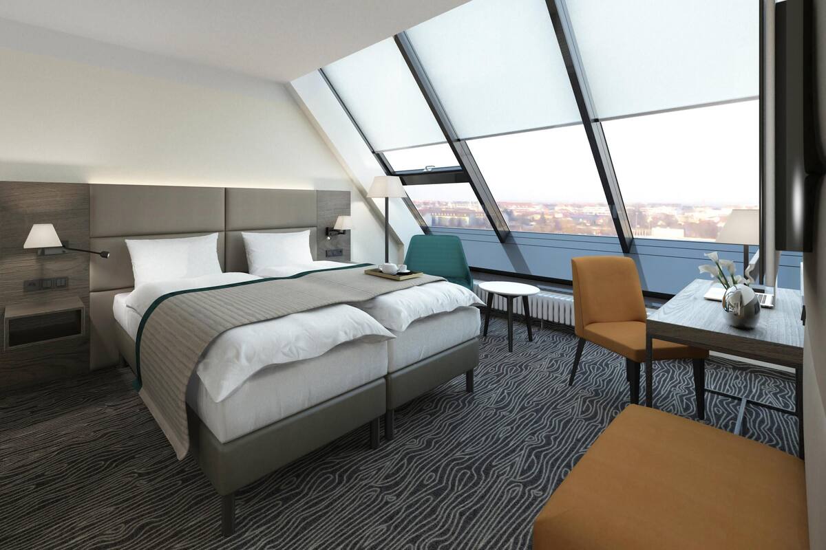 Ein modernes Hotelzimmer mit einem großen Bett, einem Schreibtisch, einem Sessel, einer Lampe und Blick auf die Stadt durch ein Oberlicht.