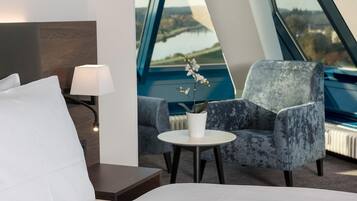 Habitación de hotel moderna con cama, una mesita de noche con lámpara, una silla y vistas a un lago a través de grandes ventanales.