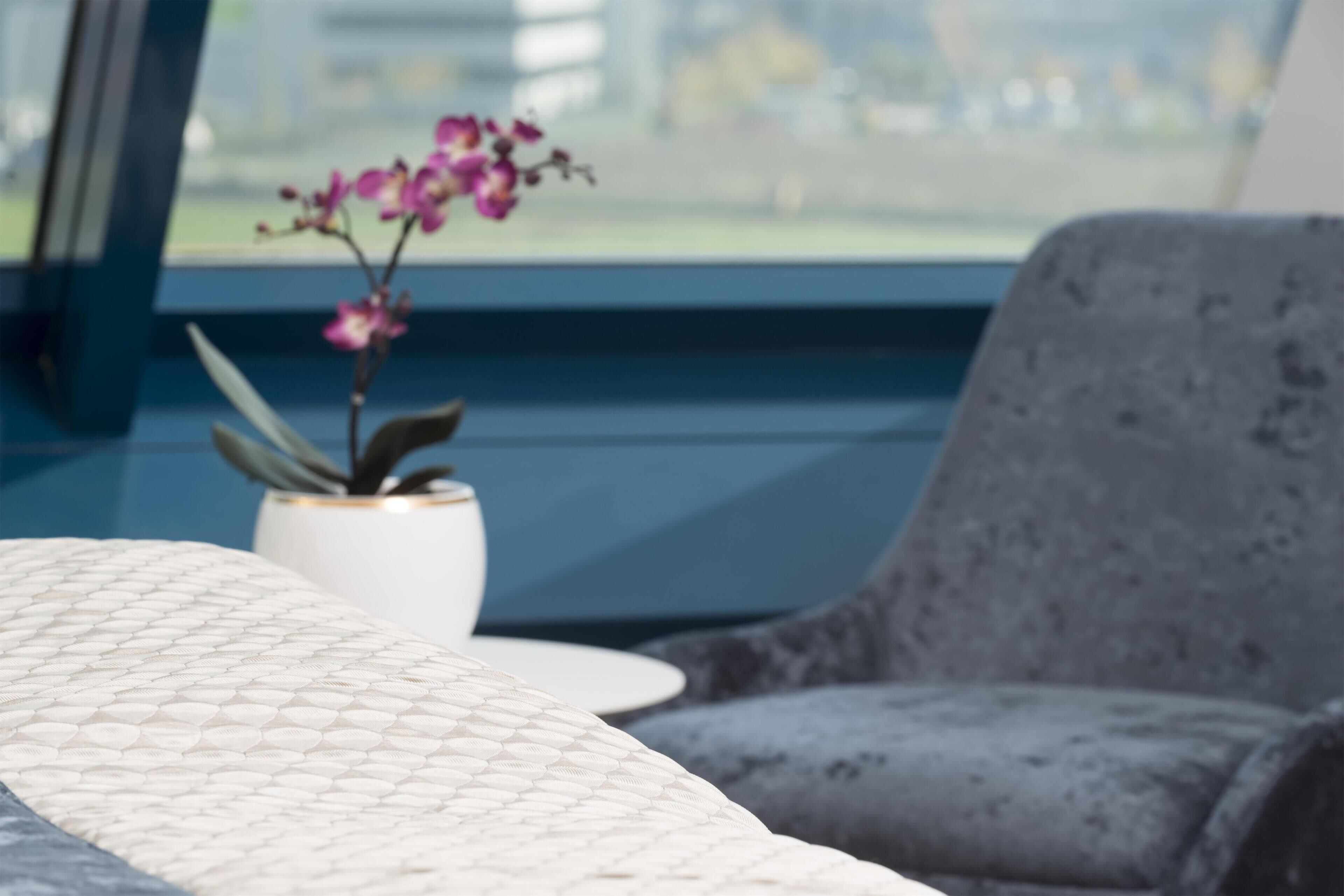 Una cama con cubre texturizado, una planta en maceta con flores moradas, un sillón tapizado gris y vistas a través de una ventana.