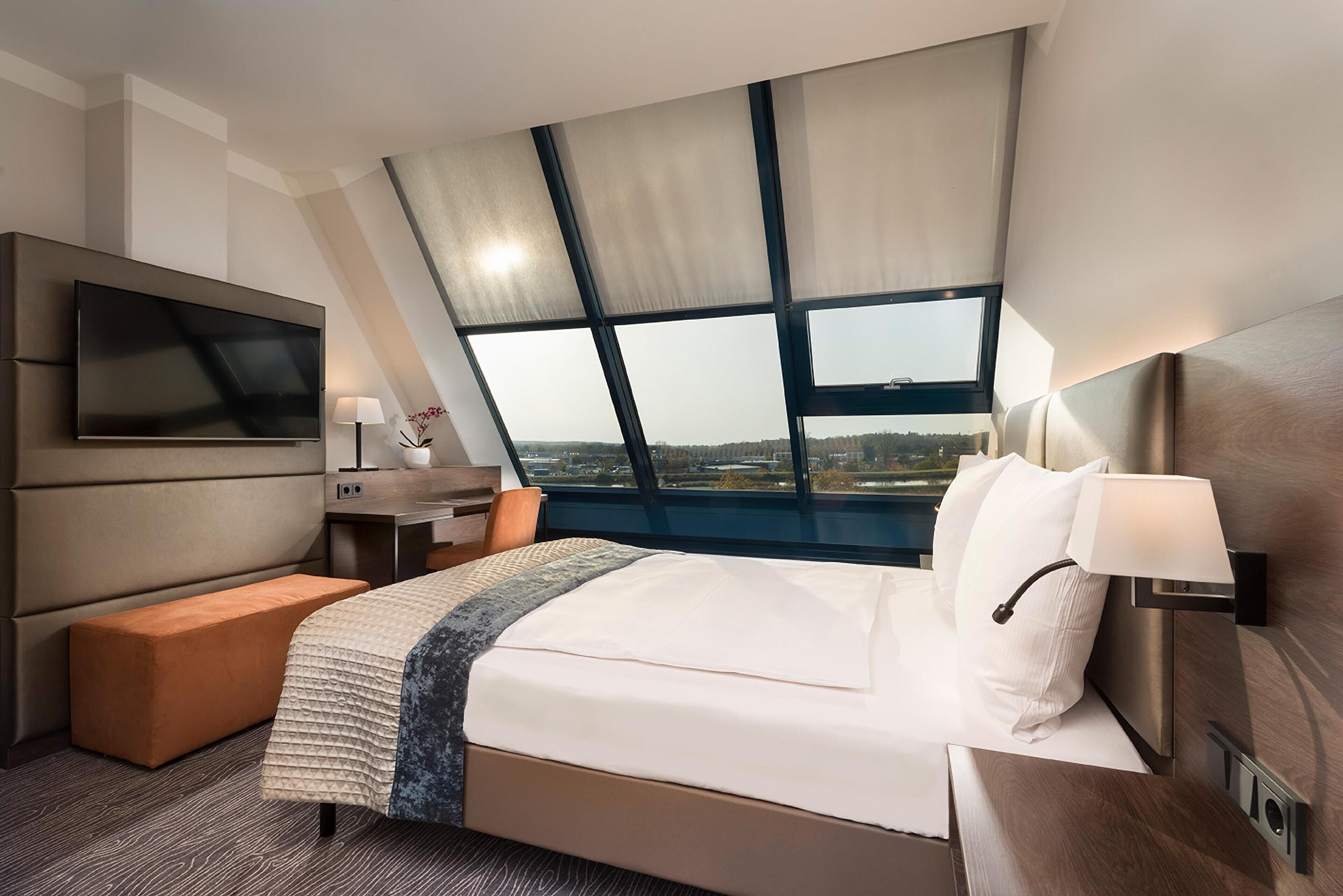 Una habitación de hotel moderna con una cama grande, un televisor de pantalla plana, un escritorio y una ventana con vistas a la ciudad.