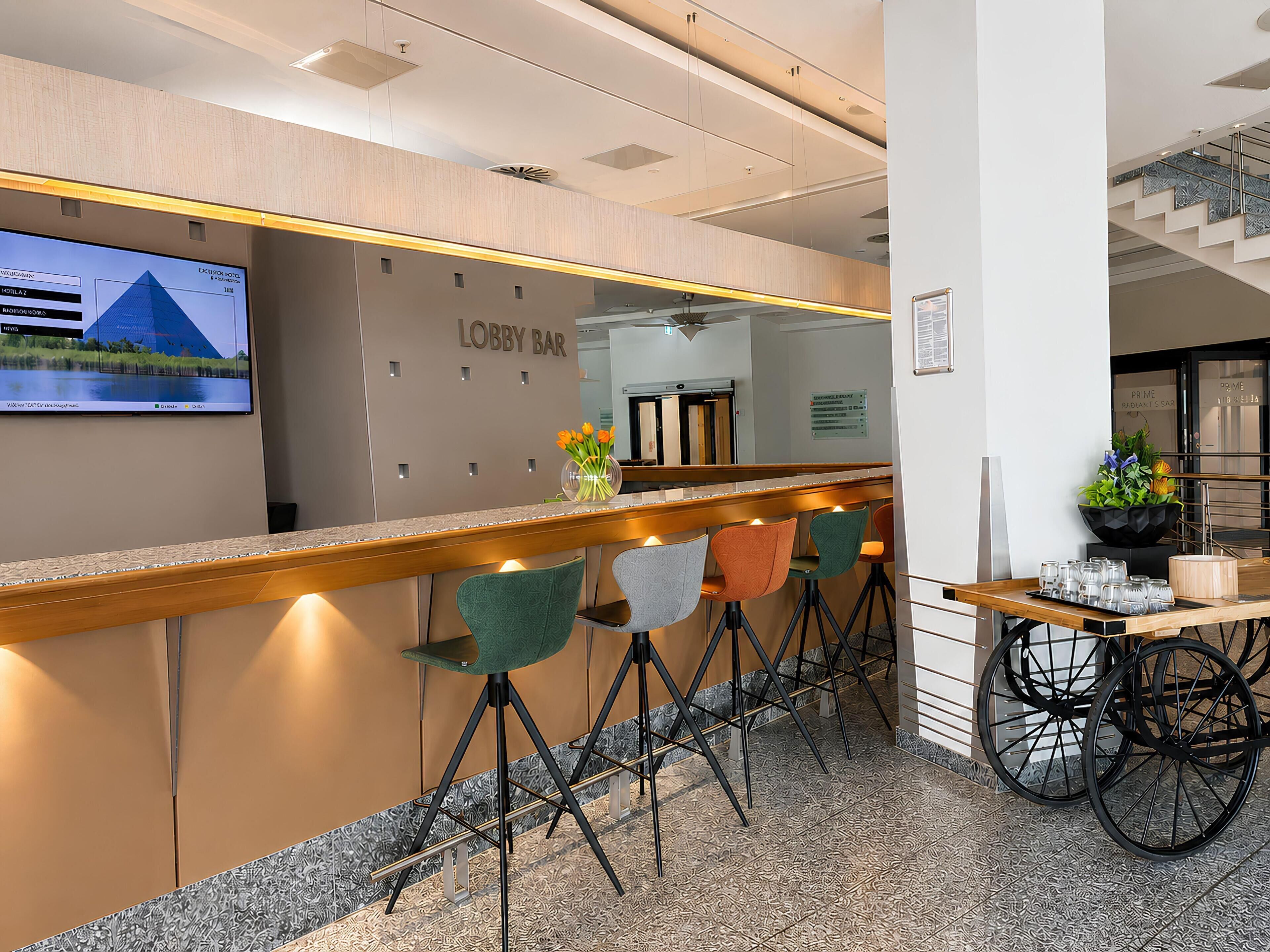 A lobby bar area with a reception desk, bar stools, and a television screen displaying a building.