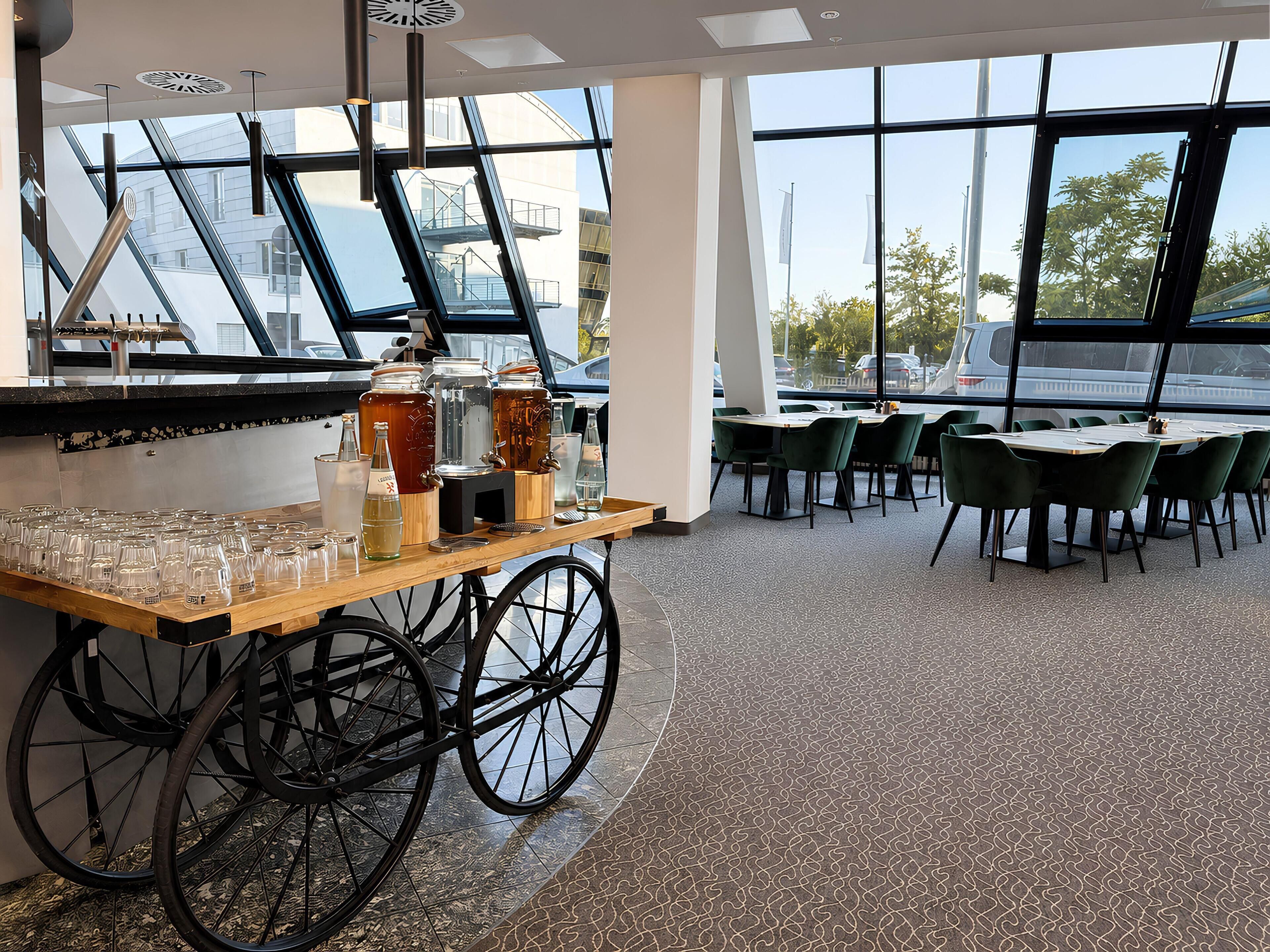 Interior de un restaurante con una zona de bar que incluye un carrito de madera con cristalería y botellas, amplias ventanas con vistas al exterior y sillas tapizadas de verde alrededor de mesas.