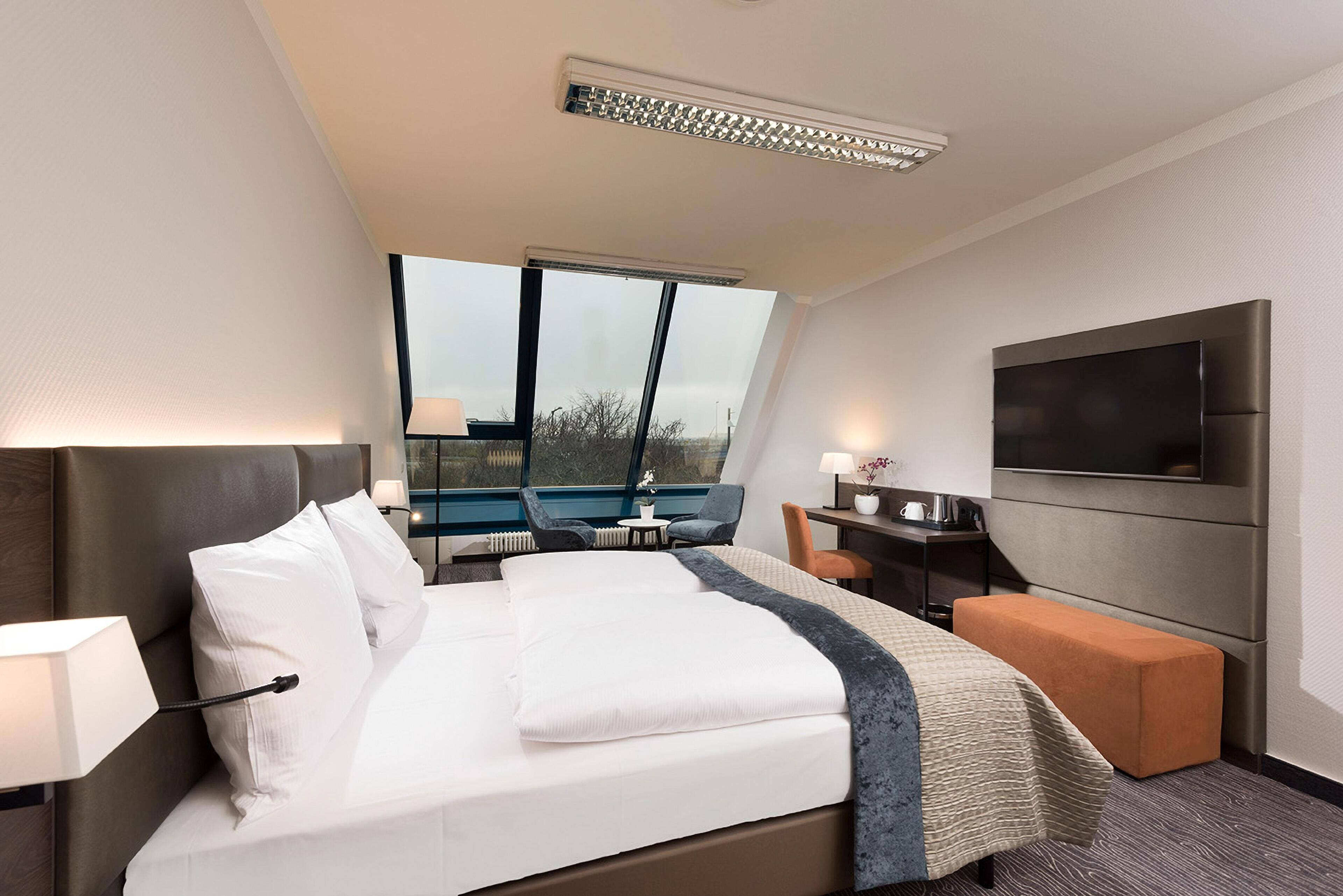 A modern hotel room with a bed, desk, chair, TV, and large windows.