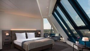 A modern hotel room with a large bed, a desk with a lamp, a chair, and a view of the city through large windows. - Excelsior Hotel Nürnberg Fürth, a member of Radisson Individuals (Fuerth)