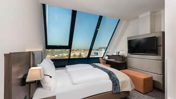 Una habitación de hotel moderna con un ventanal grande, una cama, una mesita de noche con lámpara, un televisor montado en la pared y un pequeño banco naranja.