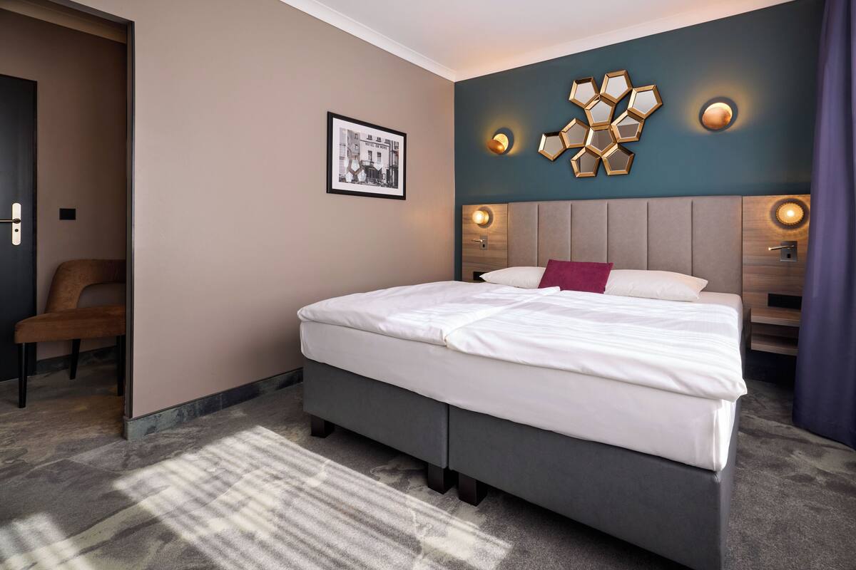 A modern hotel room with a large bed, a headboard with integrated lighting, a framed picture on the wall, and a side table with a lamp.