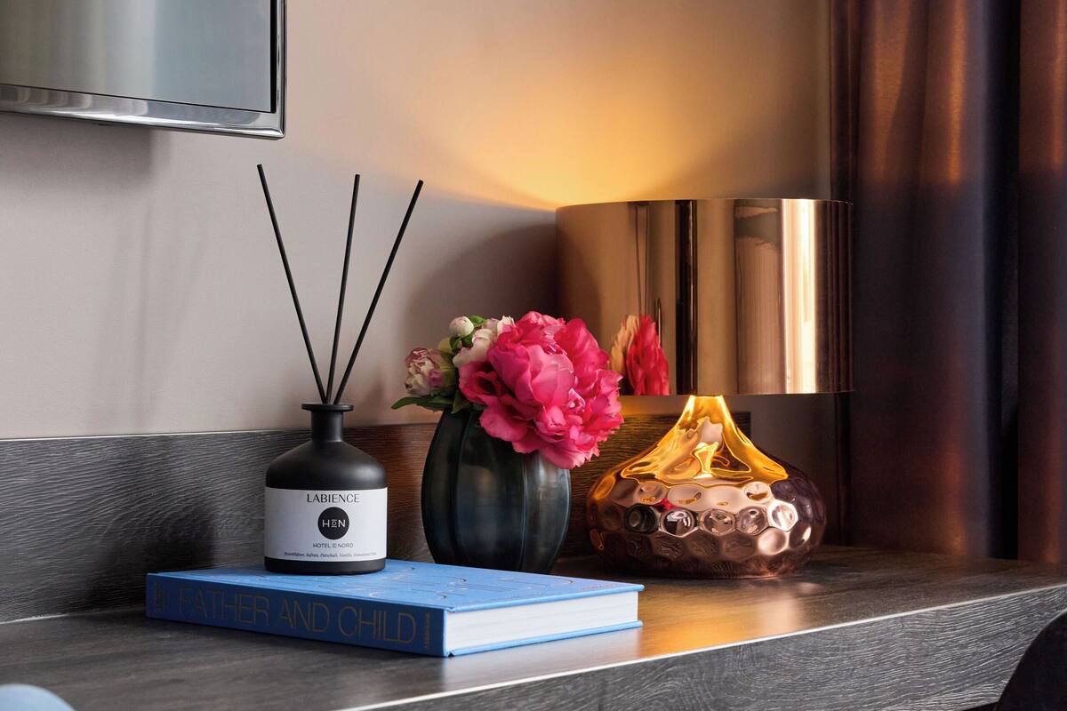 A modern hotel room with a dark wood desk, a blue book titled "Father and Child," a black vase with pink flowers, a black diffuser, and a decorative glass object.