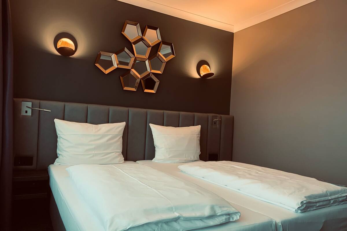 A double bed with white linens, a grey upholstered headboard, and two wall-mounted sconces.