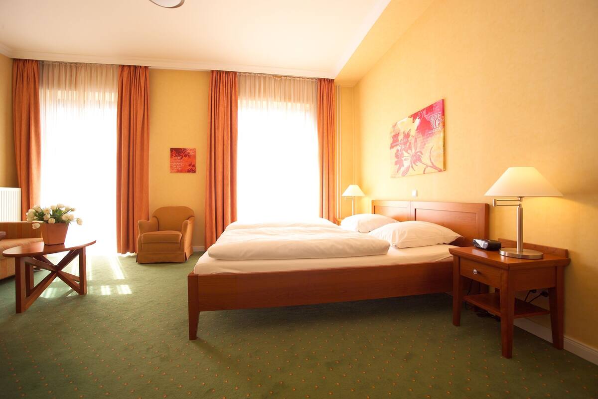A hotel room with a bed, two bedside lamps, a chair, a small table with a plant, and a painting on the wall.