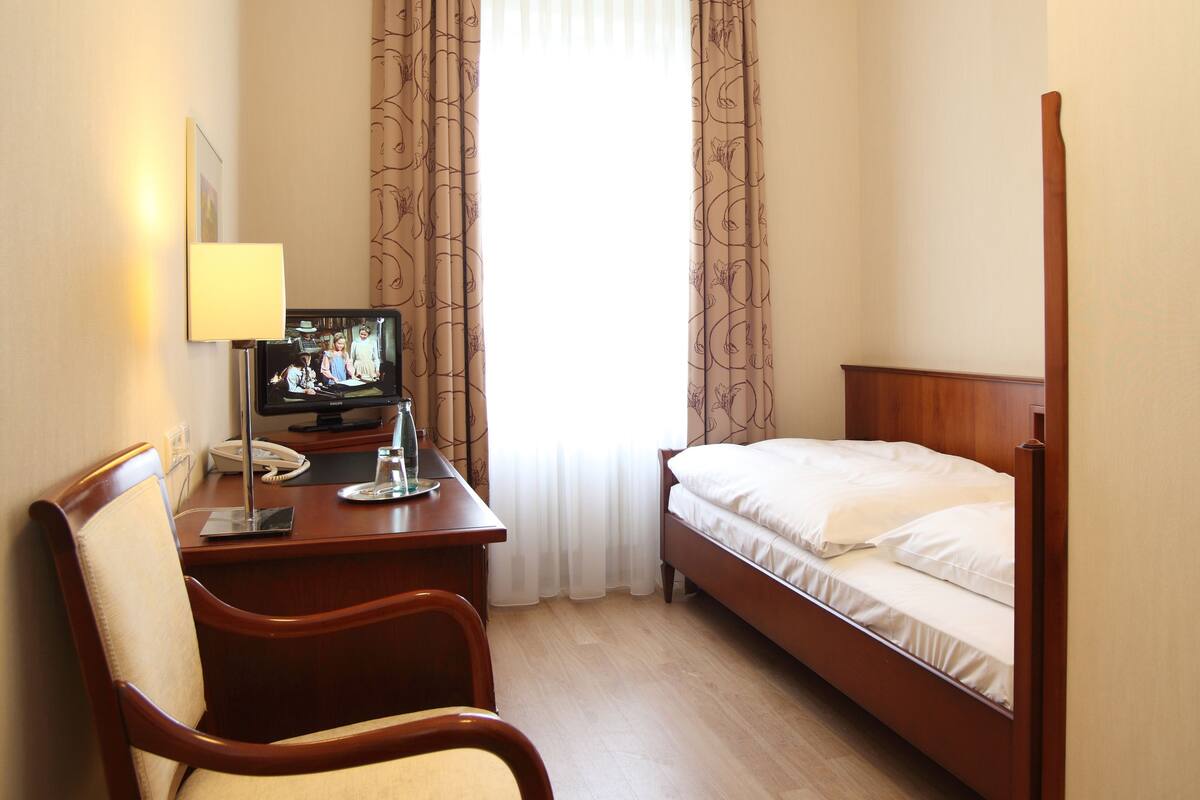 A hotel room with a bed, a desk with a chair, a television, and a window with curtains.