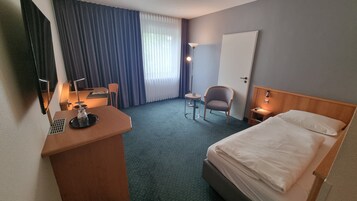 Ein Hotelzimmer mit einem Bett, einem Schreibtisch mit Stuhl, einem kleinen Tisch, einer Lampe und einem Fenster mit Vorhängen.