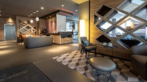 A modern lobby with a checkered floor, a seating area with a sofa and chairs, a fireplace, and a kitchen area visible through a large window. - Hotel Panorama Harburg (Hamburg)