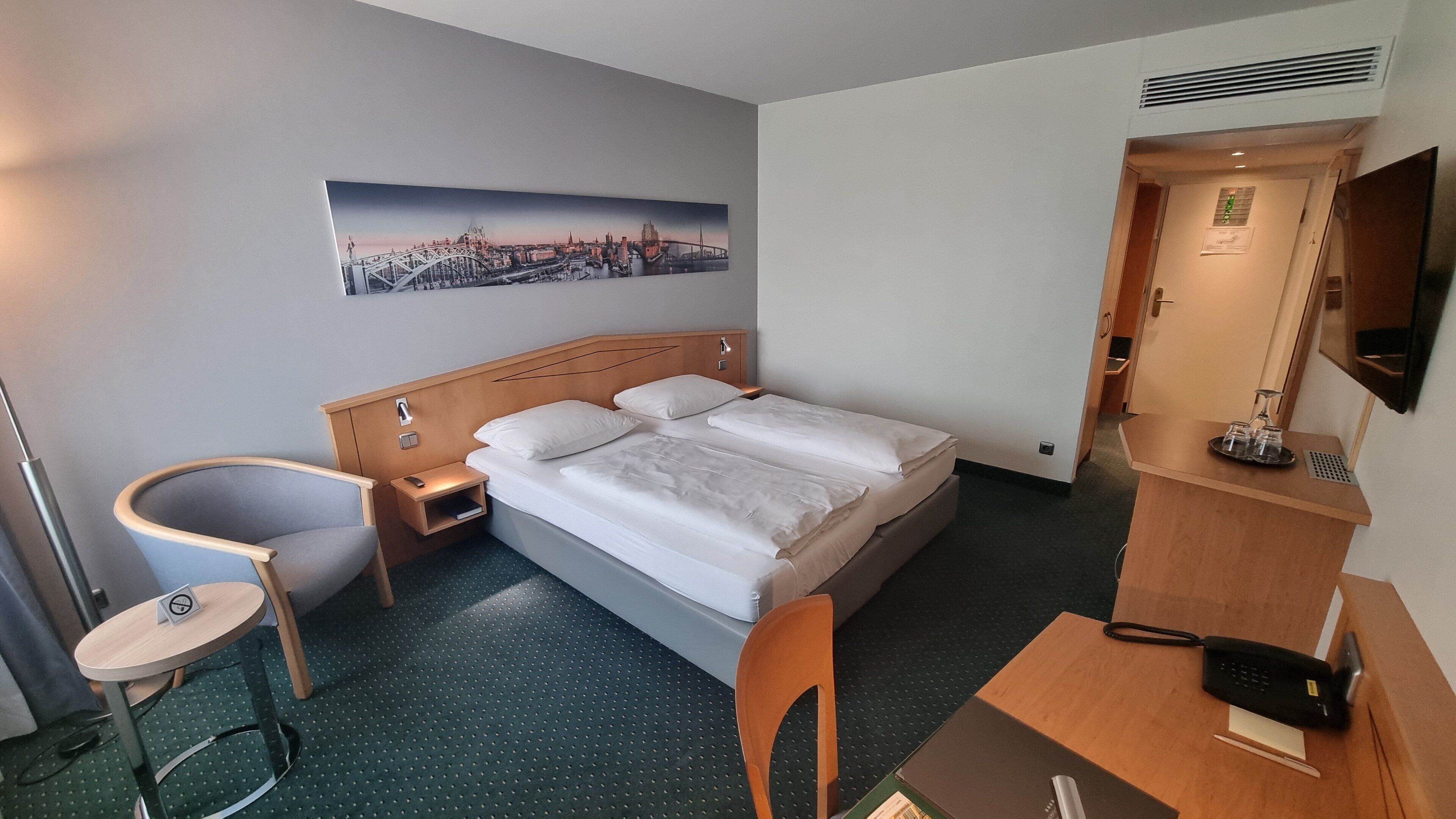 Habitación de hotel con cama, escritorio, silla, mesita, televisor y un cuadro en la pared.