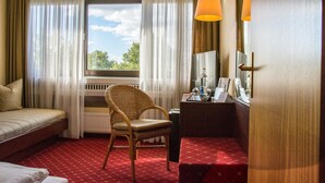 A hotel room with a bed, a chair, a desk with a lamp, a window with curtains, and a view of trees. - Hotel Helgoland (Hamburg)