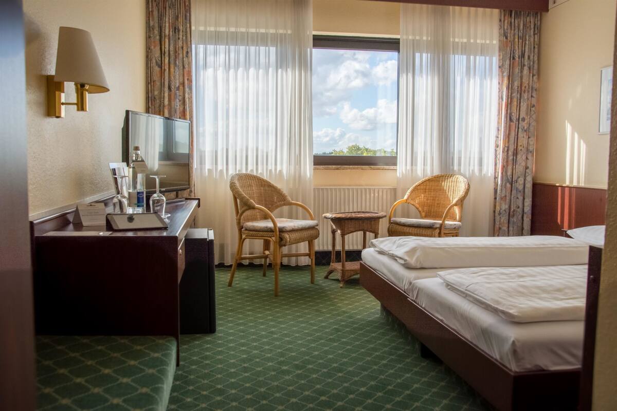 A hotel room with a bed, two wicker chairs, a small table, a desk with a lamp, and a television.