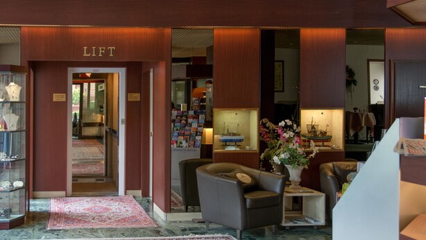 A lobby with a reception desk, a seating area with a brown leather sofa, a coffee table, and a vase with flowers. There is a display of postcards and a small shelf with decorative items. - Hotel Helgoland (Hamburg)