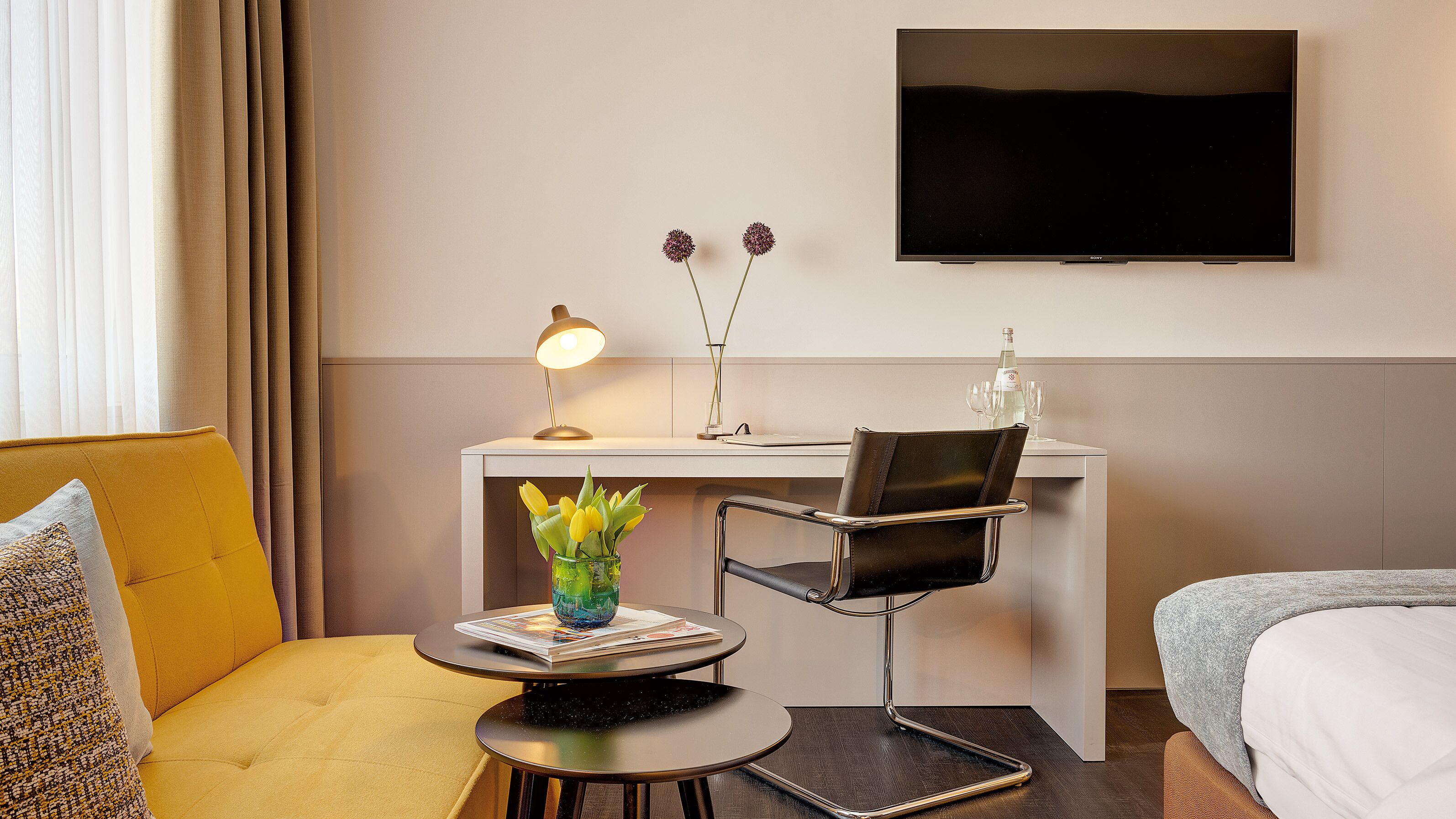 Habitación de hotel con una cama, un escritorio con una silla, una lámpara, un florero con flores y un televisor de pantalla plana en la pared.
