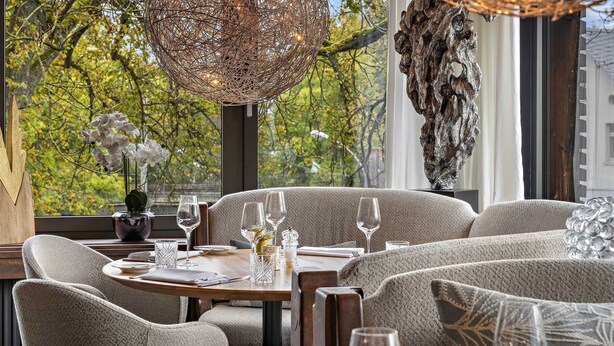 Un comedor con una mesa redonda preparada para cuatro personas, rodeada de sillas tapizadas. Sobre ella, un candelabro metálico decorativo, y a la derecha se observa una gran escultura abstracta.