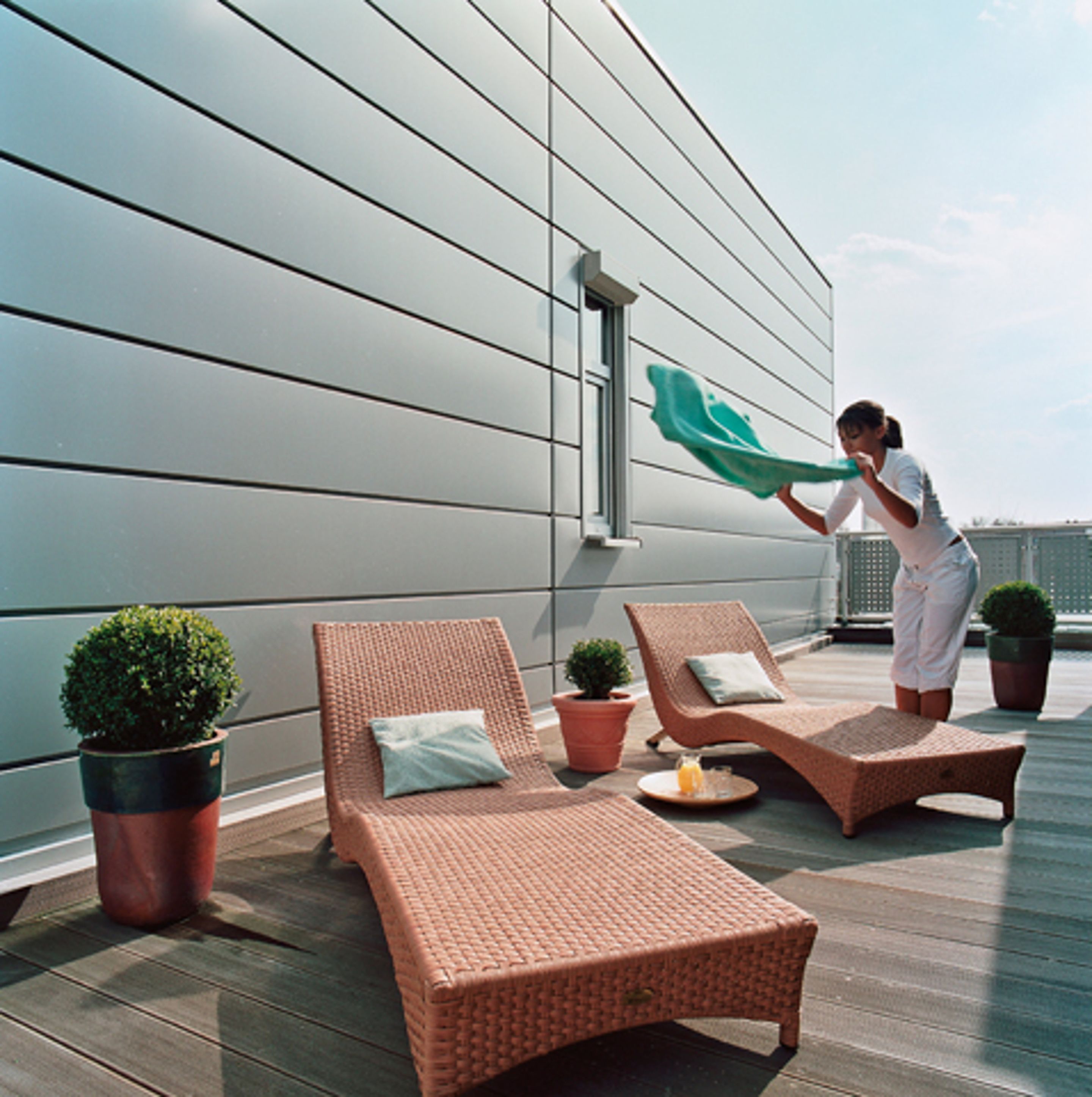 Een terras op het dak met rieten lounge fauteuils, planten in potten en iemand die een handdoek opvouwt.