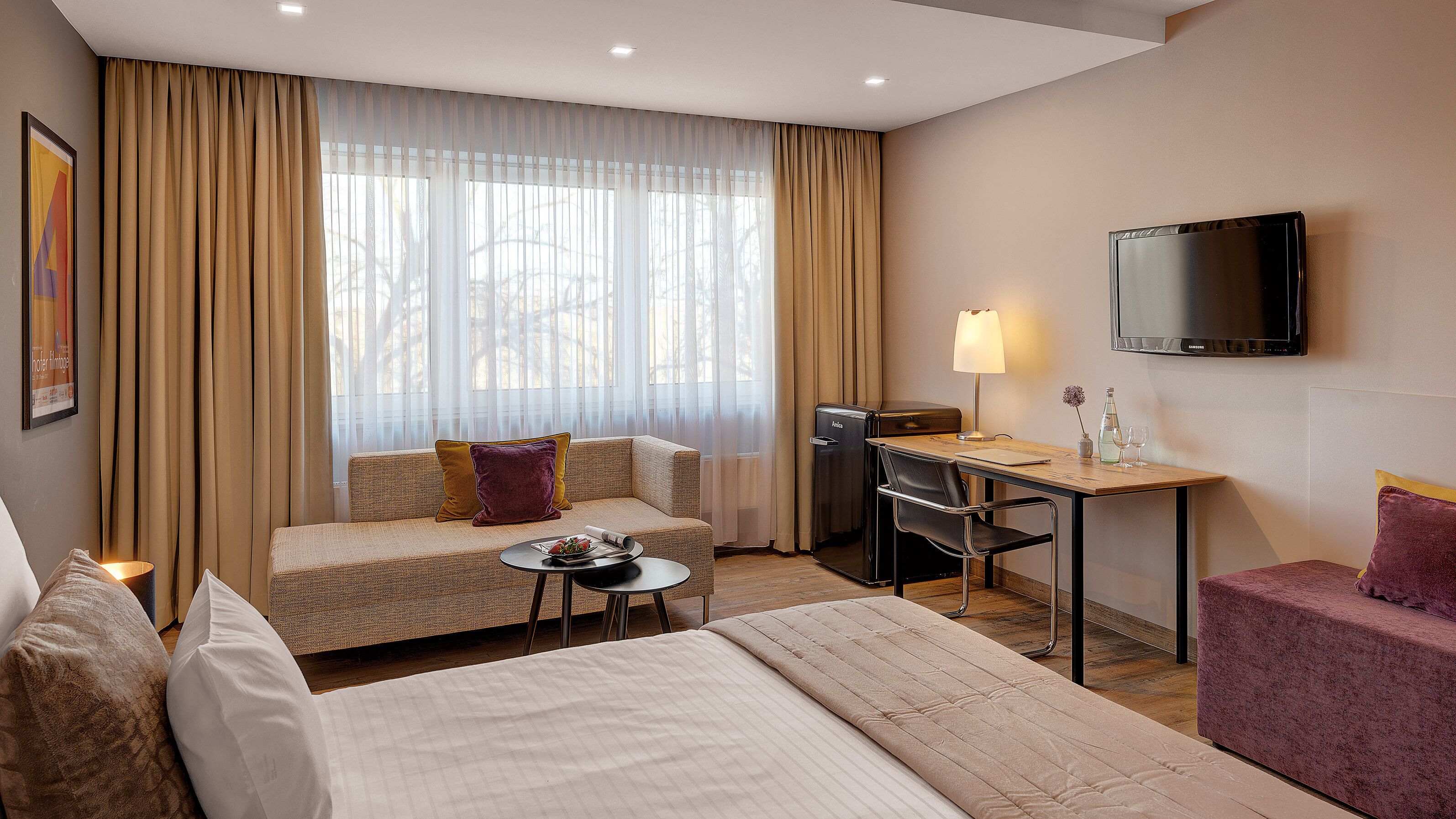 A hotel room with a bed, a sofa, a desk with a chair, a TV, and a minibar.