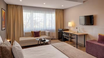 A hotel room with a bed, a sofa, a desk with a chair, a TV, and a minibar.