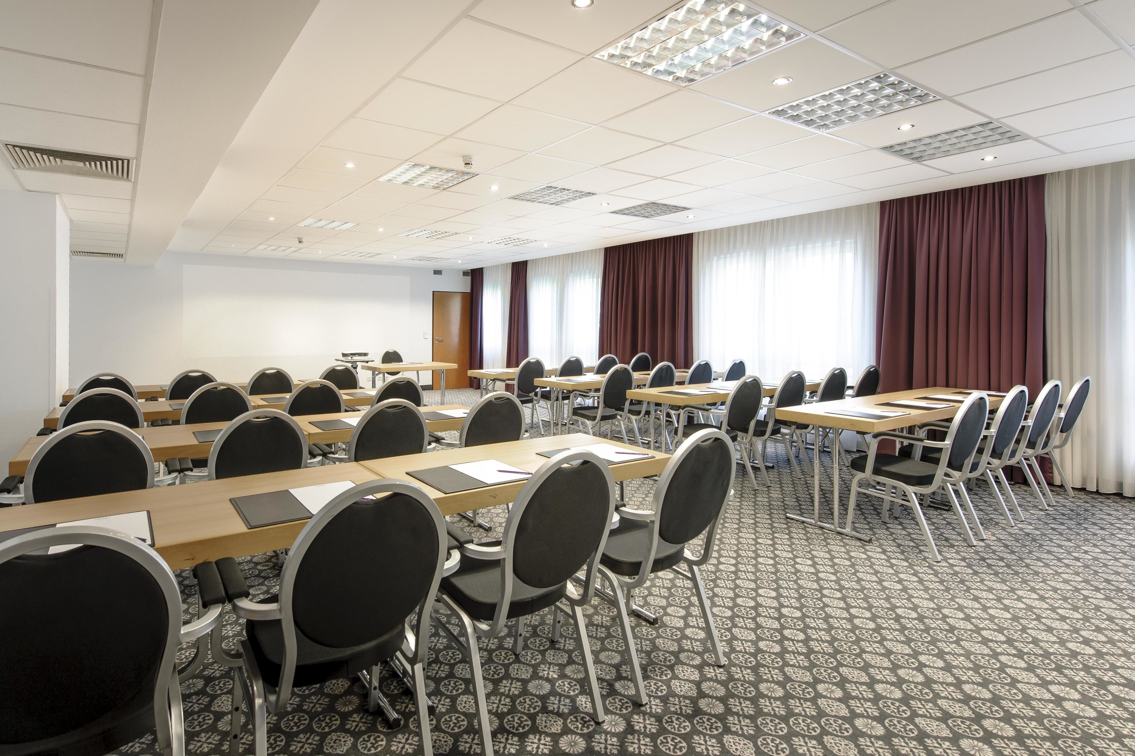 A conference room with multiple tables and chairs, arranged in rows, with a patterned carpet and a large window with curtains.