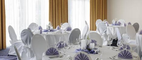 Un salón de banquetes con mesas redondas dispuestas para comer, sillas blancas y servilletas moradas.