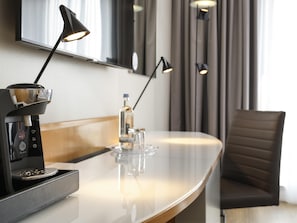 Quarto de hotel moderno com cafeteira, mesa de vidro, cadeira e espelho.