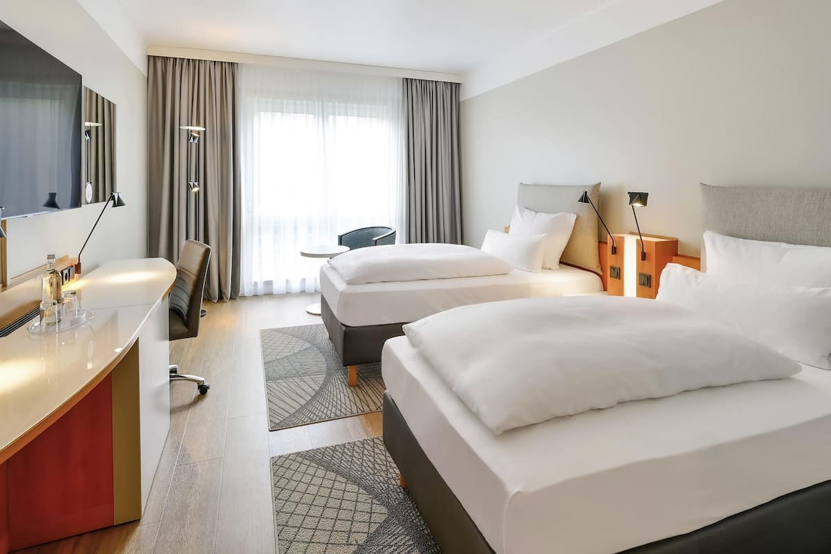 Quarto de hotel com duas camas, uma escrivaninha, uma cadeira, uma televisão e uma janela com cortinas.