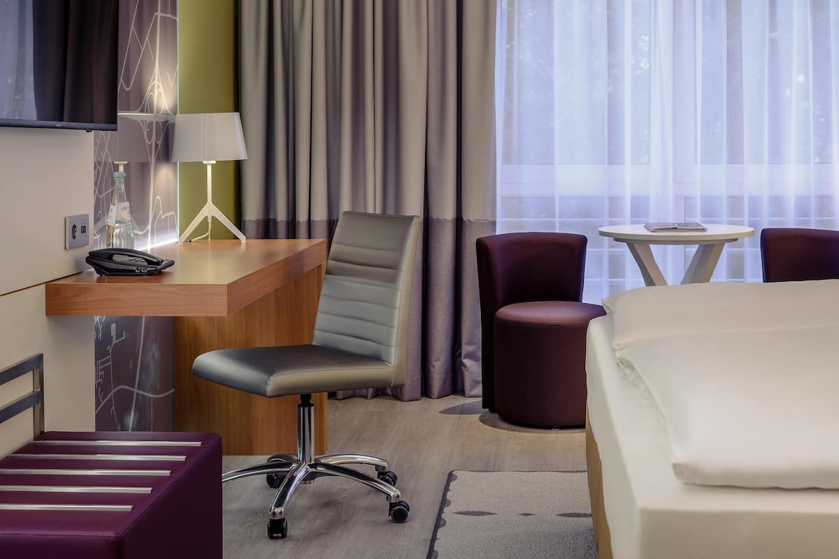 Quarto de hotel com escrivaninha, uma cadeira, uma cama, uma mesinha e janela com cortinas.