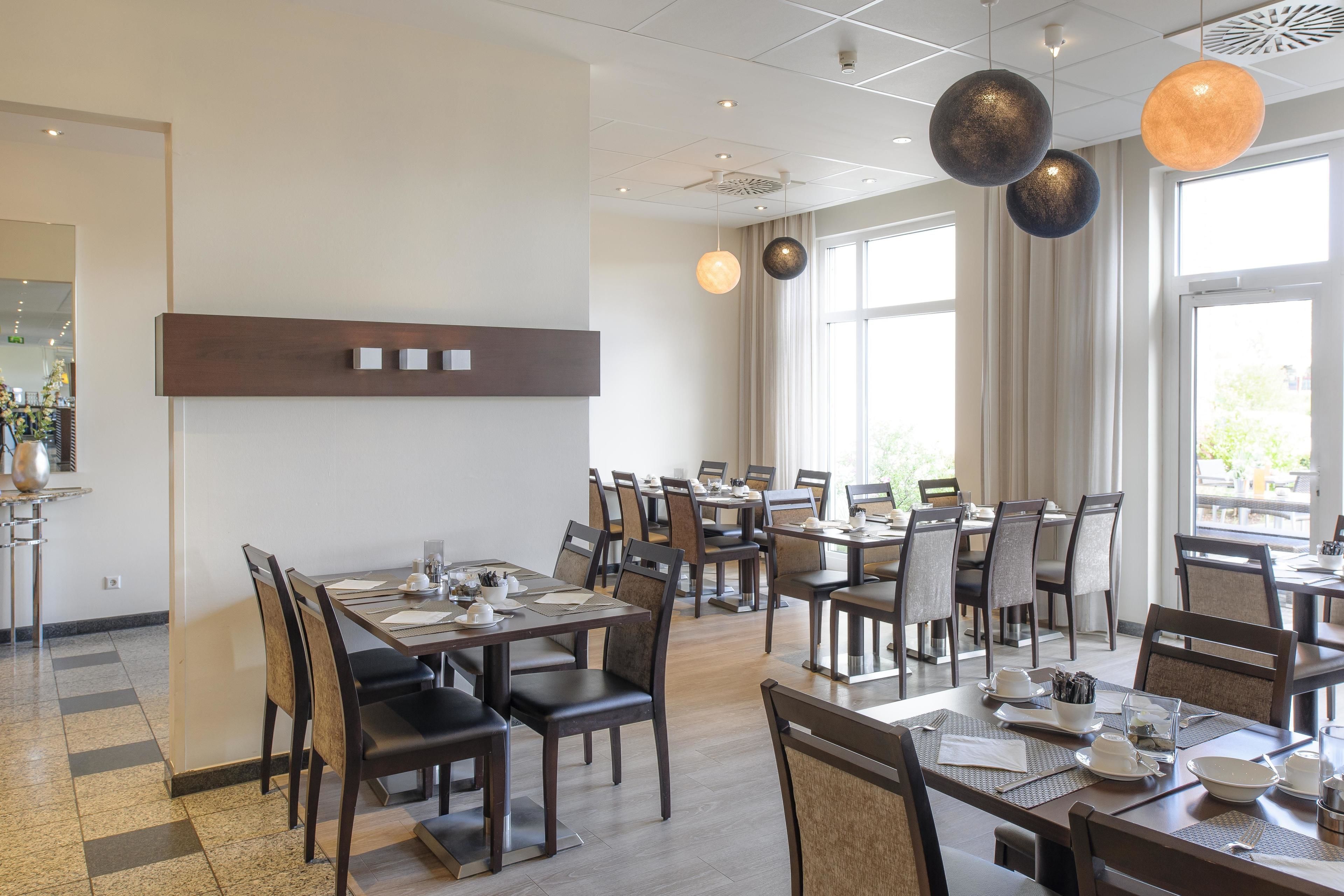 A spacious dining area with multiple tables and chairs, large windows allowing natural light, and modern pendant lighting fixtures.