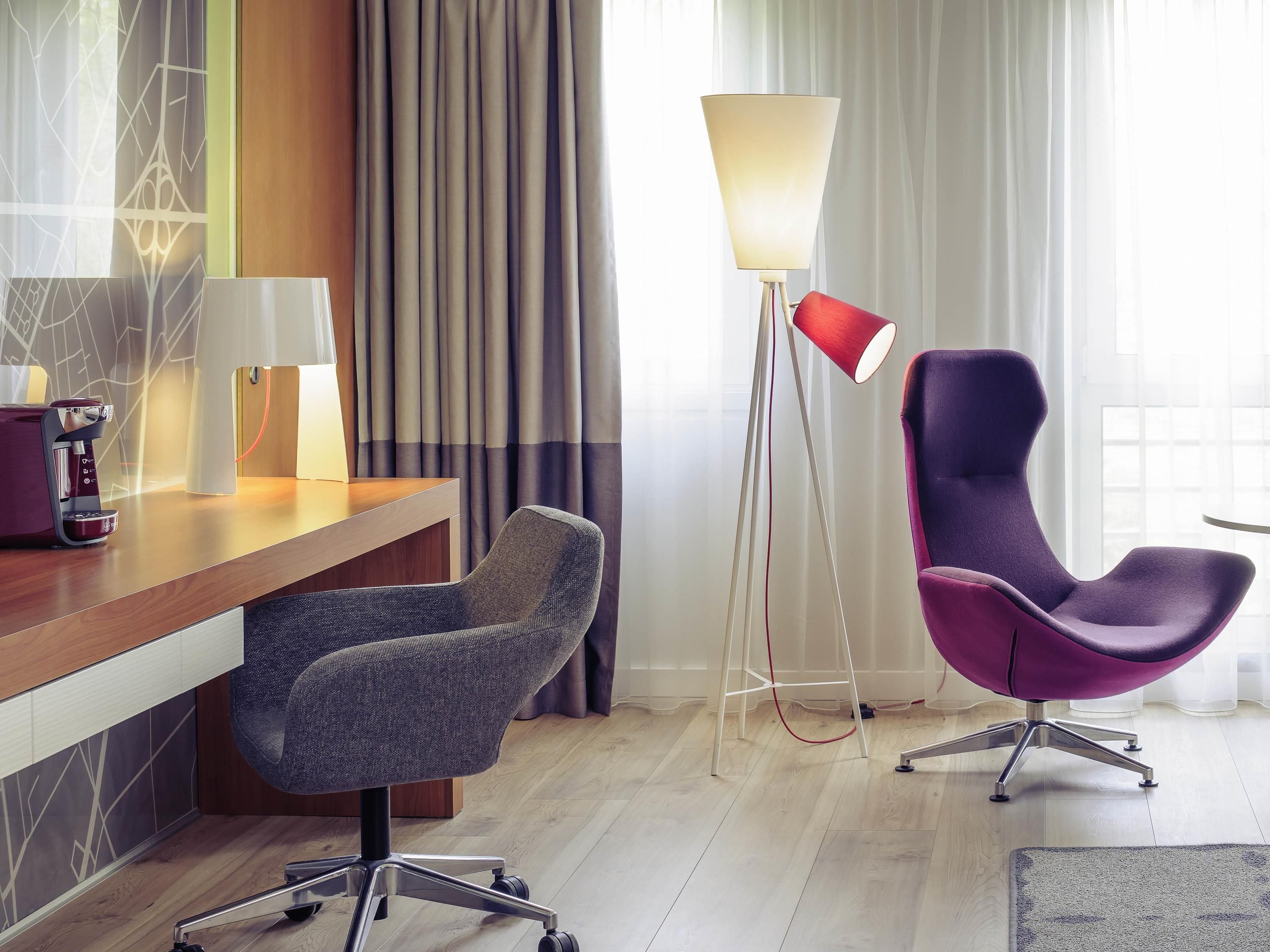 A modern hotel room with a wooden desk, a purple chair, a standing lamp, and a large window with curtains.