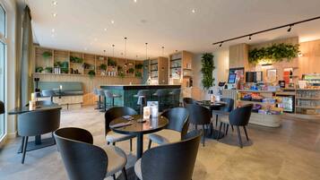 Ein modernes Café mit runden Tischen und Stühlen, einer Bar und Regalen mit Pflanzen und Getränken.