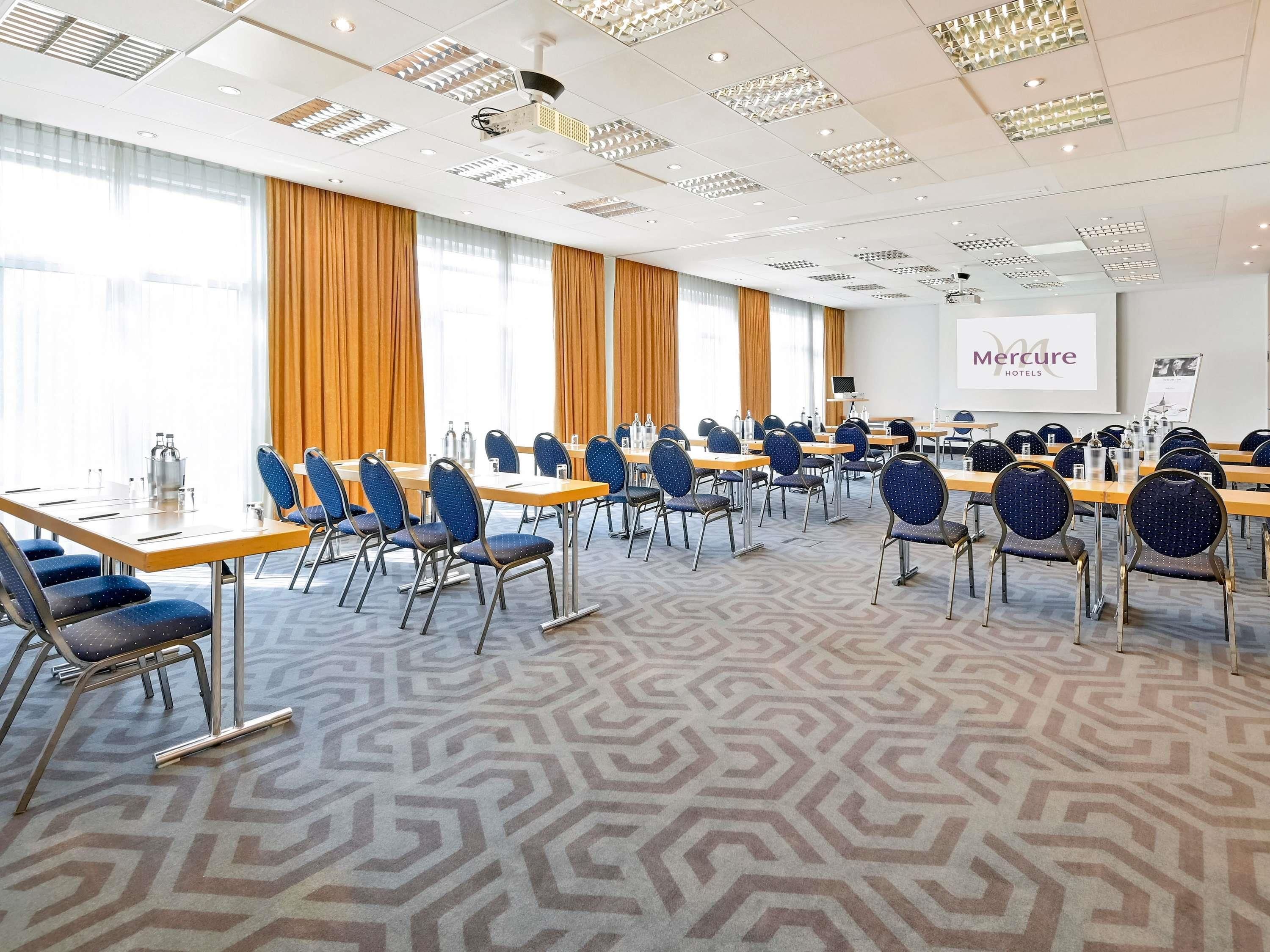 A conference room with rows of tables and chairs, a projector screen, and a large window with curtains.