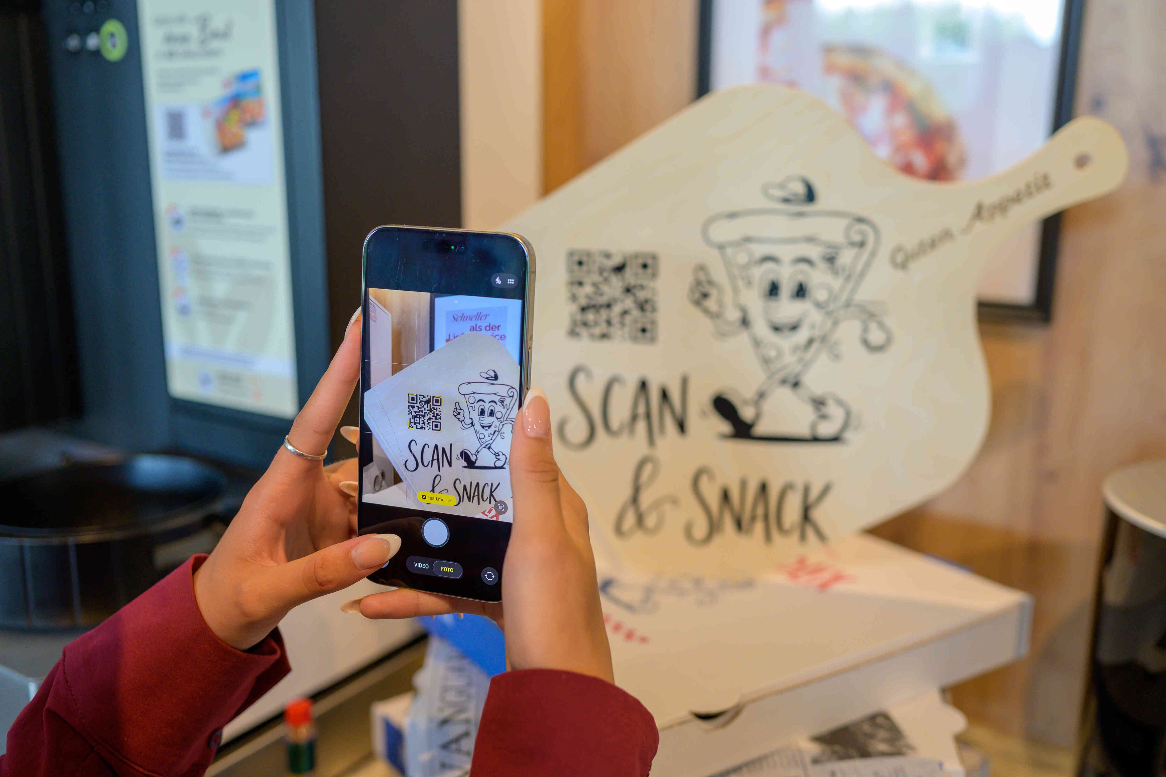 A person holding a smartphone with a QR code and a sign that reads 'SCAN & SNACK' with a cartoon character.