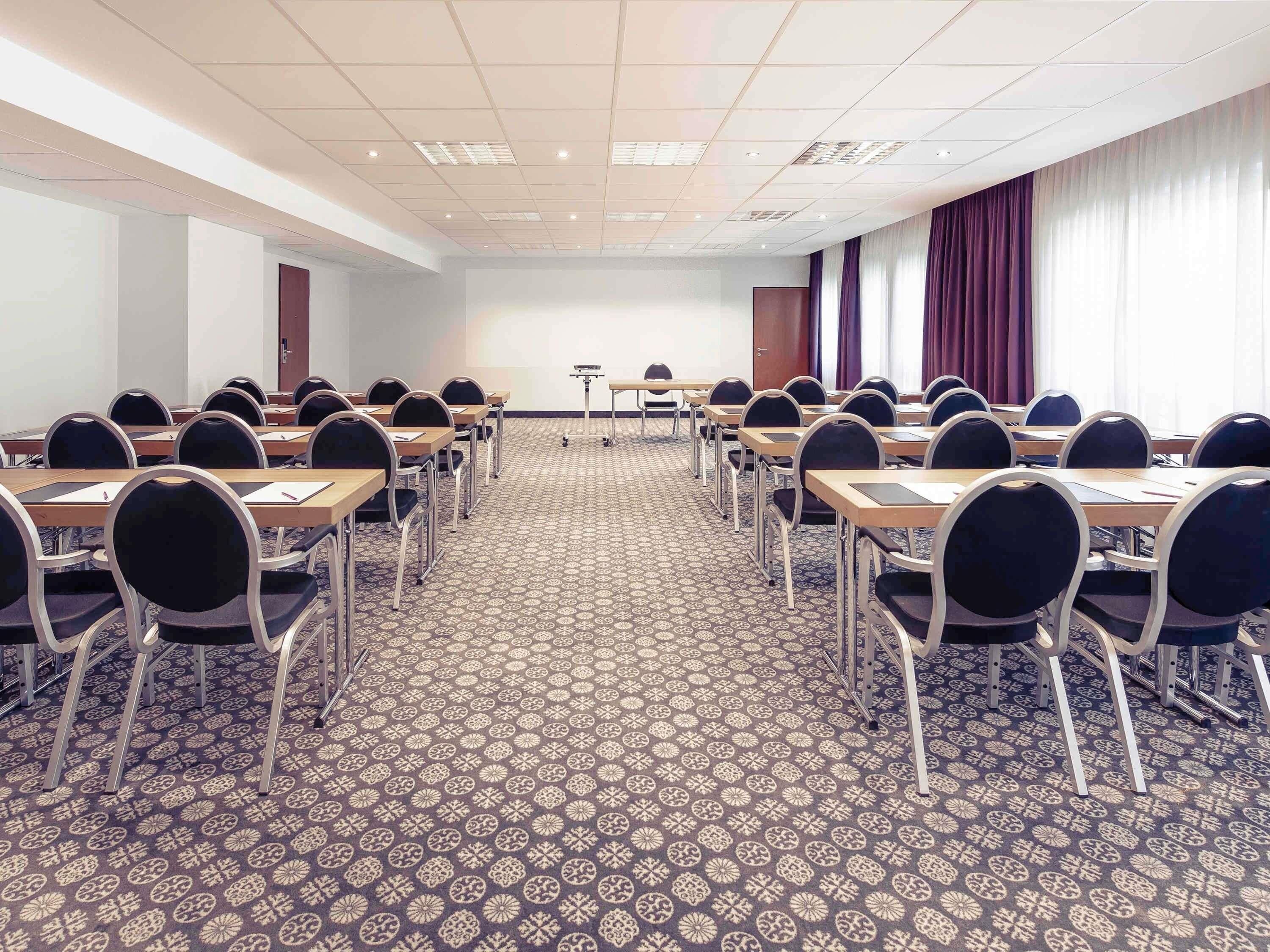 A conference room with rows of tables and chairs, a projector screen, and a patterned carpet.