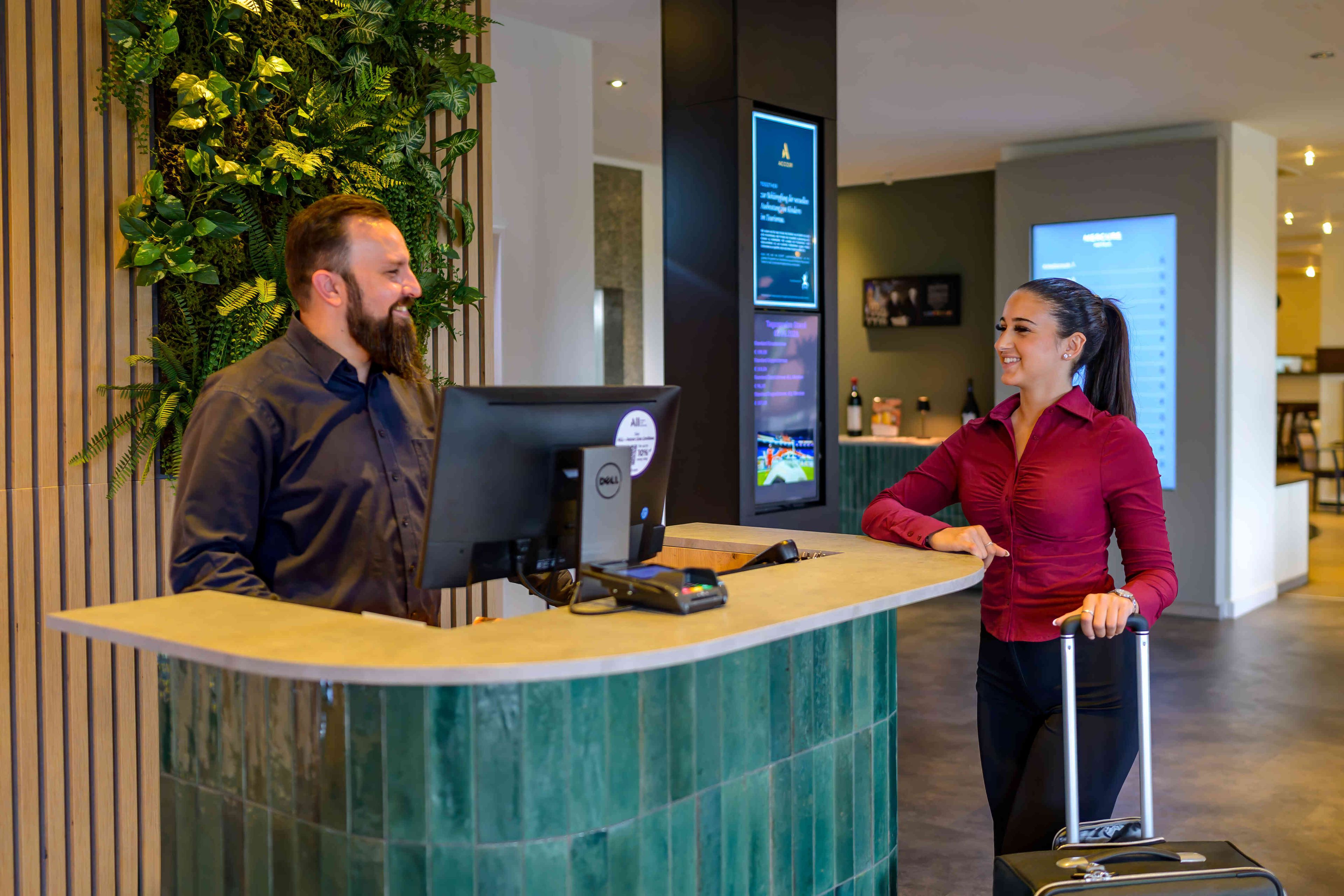 Un mostrador de recepción con una computadora, una pantalla digital que muestra información y una pared decorada con plantas verdes.