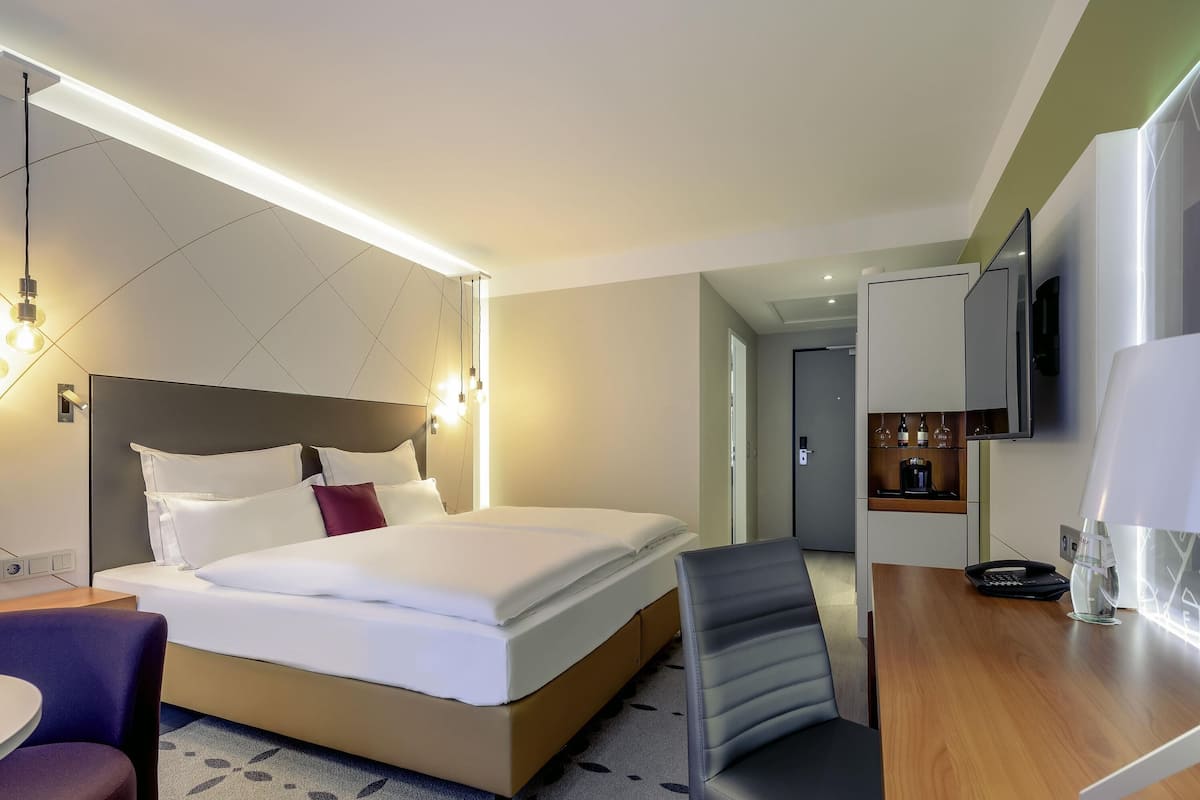 Quarto de hotel moderno com cama, escrivaninha, cadeira, televisão e minibar.