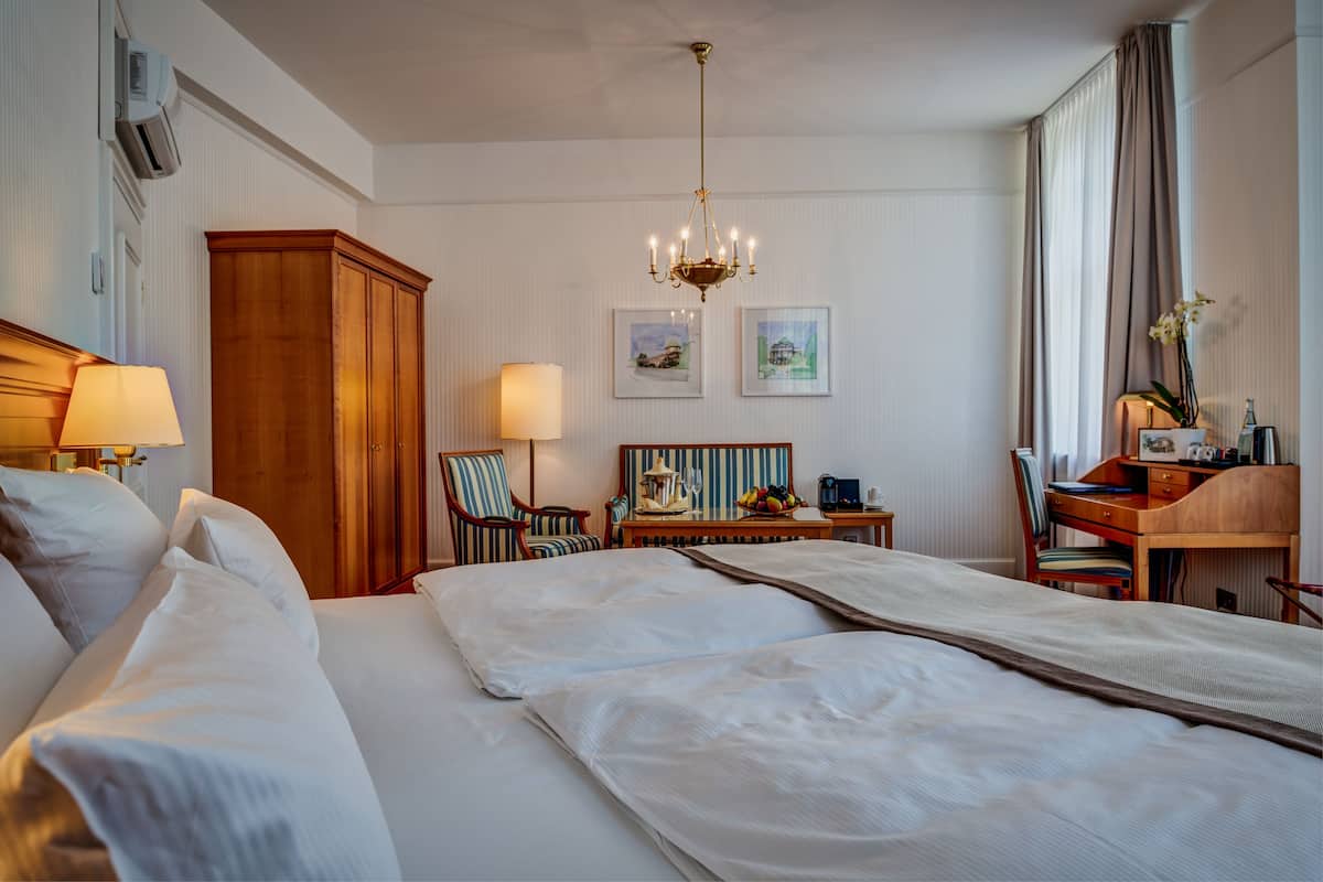 A hotel room with a large bed, a desk, a chair, a lamp, and two framed pictures on the wall.