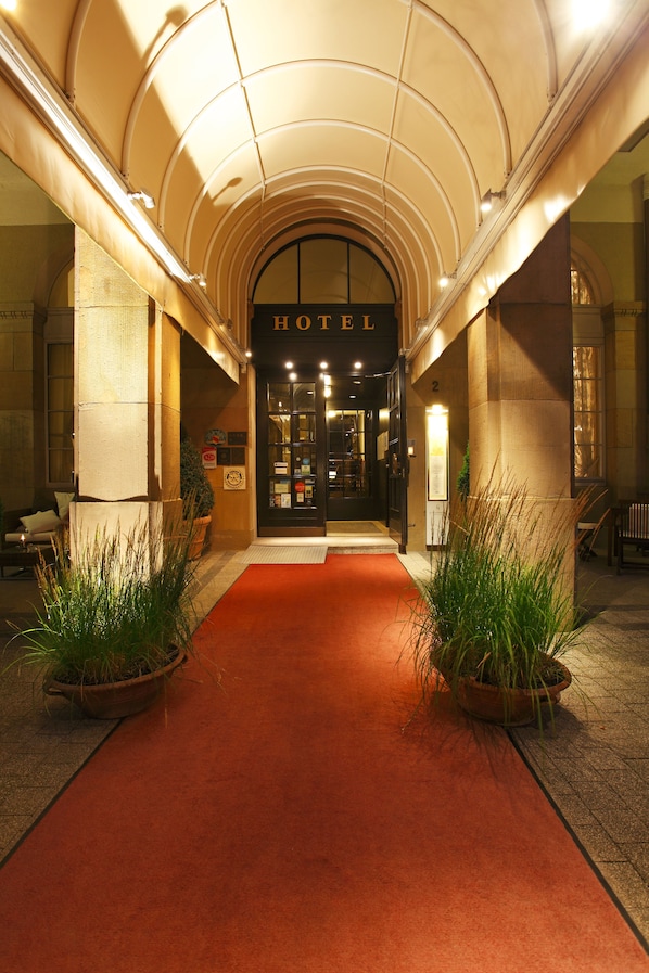 A hotel entrance with a red carpet, arched ceiling, and potted plants on either side.