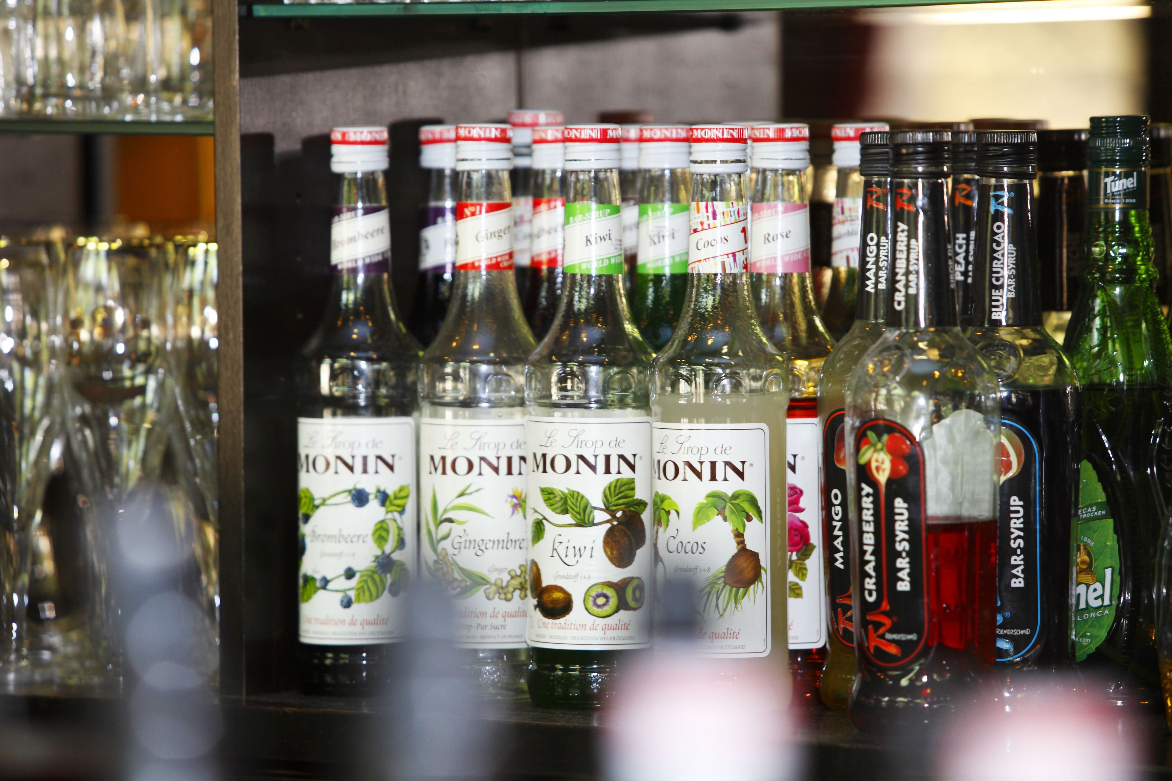Una repisa con varias botellas de sirope Monin, incluyendo sabores como jengibre, kiwi y mango.