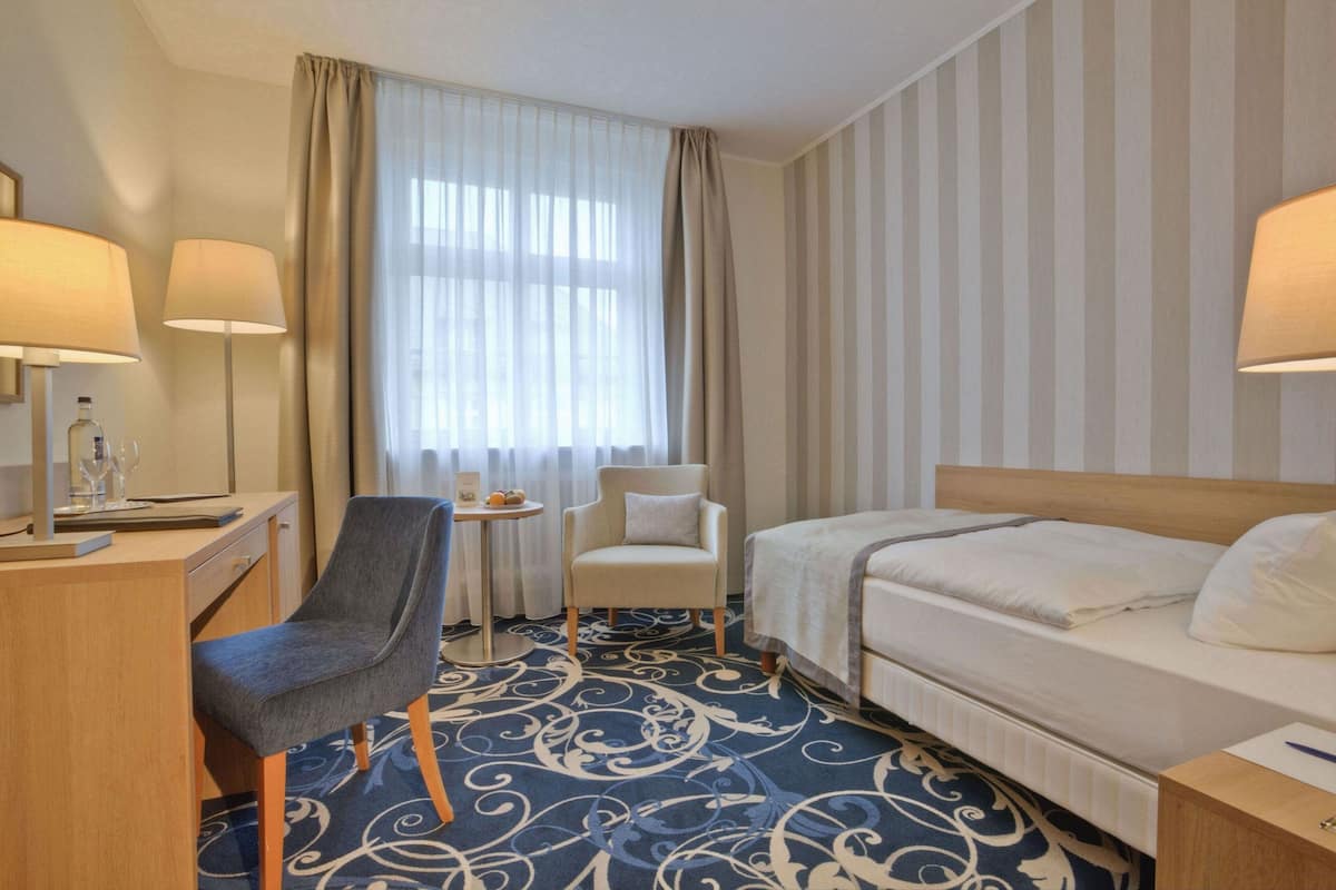 A hotel room with a bed, a desk with a lamp, a chair, a small table, and a window with curtains.