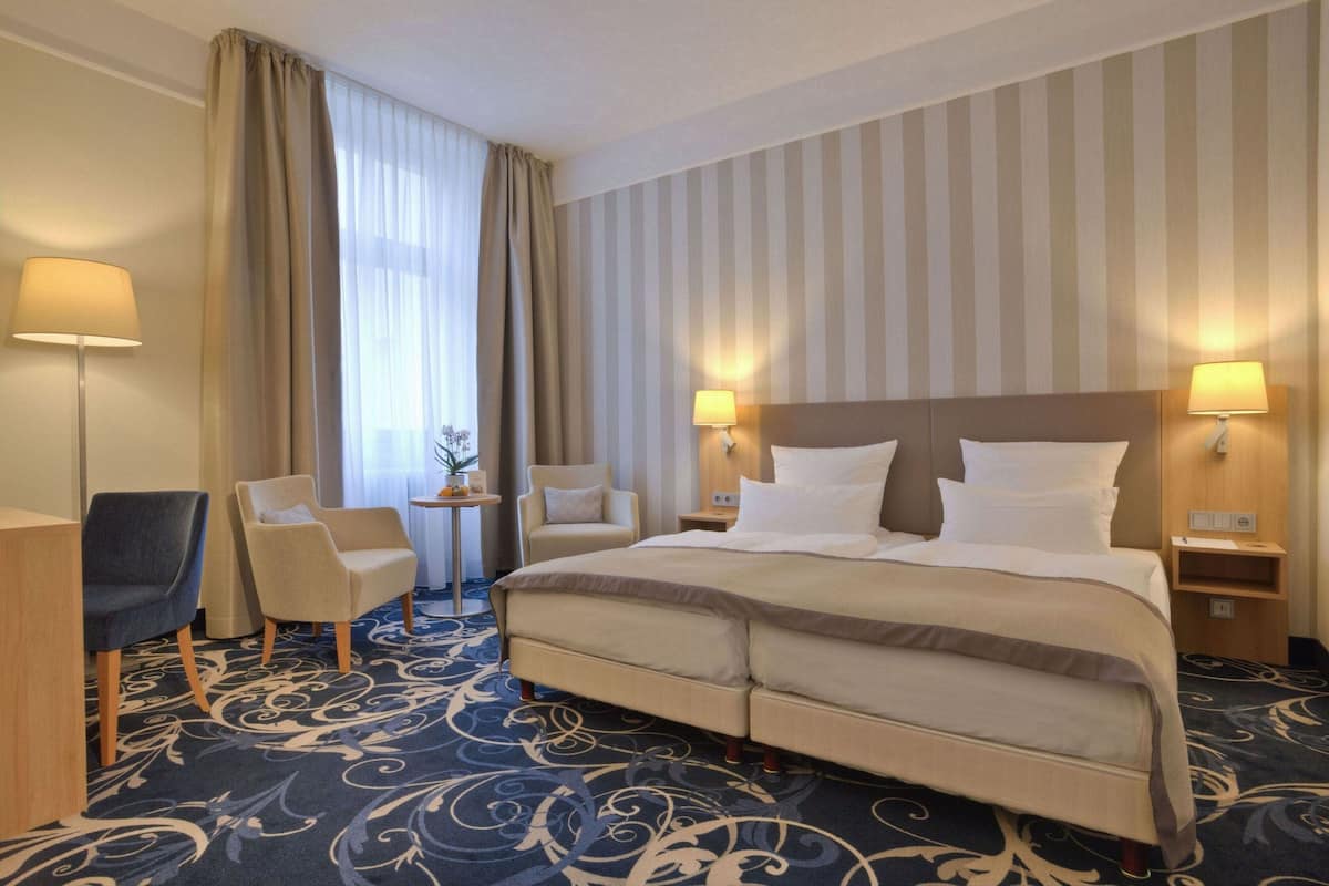 A hotel room with a large bed, two bedside tables with lamps, a chair, a small table with a plant, and a window with curtains.