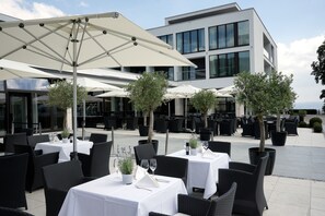 Tables à manger en plein air, équipées de parasols, chaises en osier noir et plantes en pot.